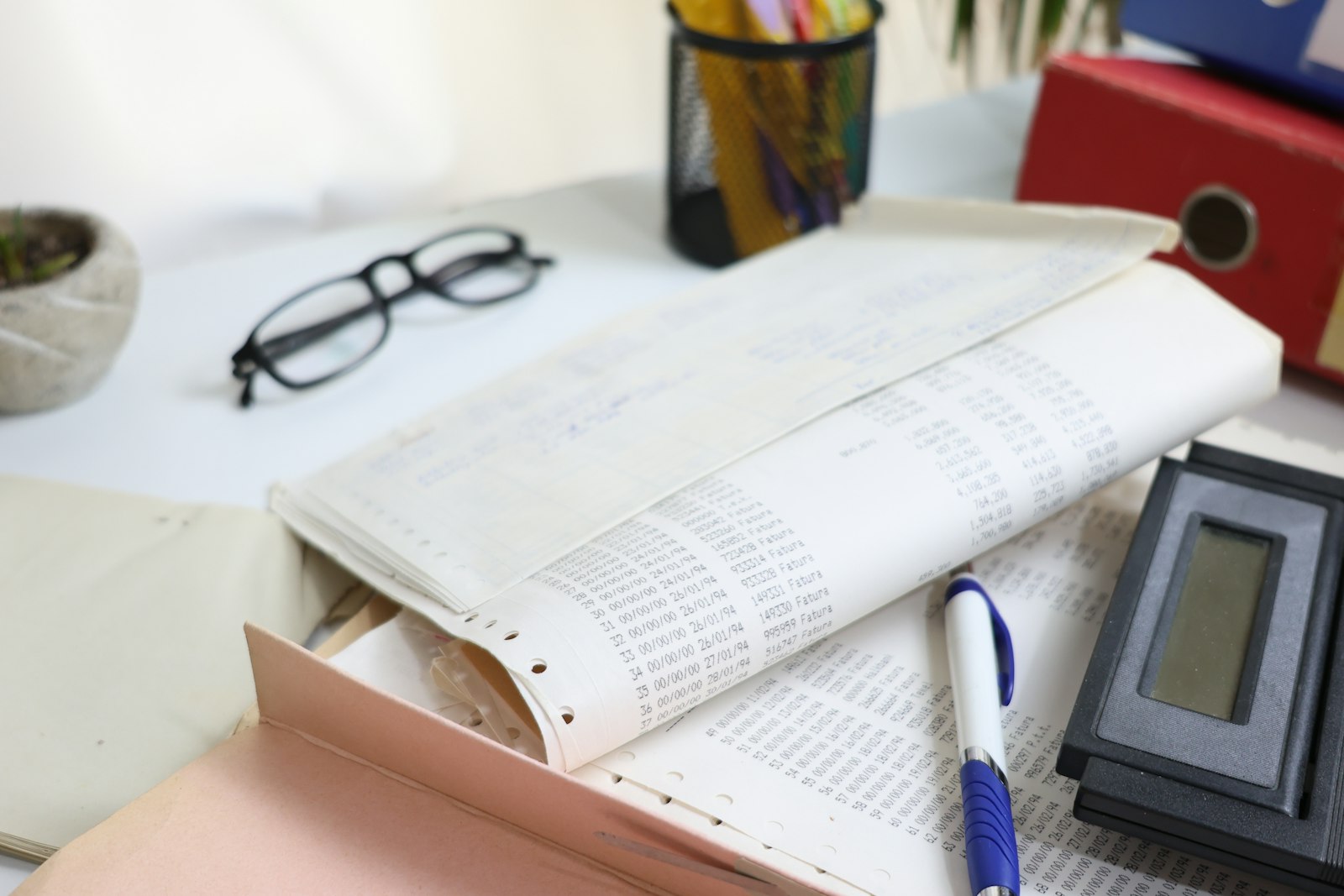 A stack of old dot-matrix invoice printouts next to a calculator and reading glasses on a desk